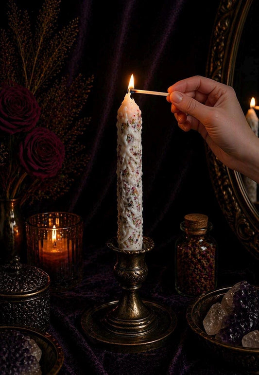 May include: A white candle with a textured design and colorful speckles is lit with a match. The candle sits in a vintage brass candlestick. Other items on the table include a lit candle, a jar, and a bowl.